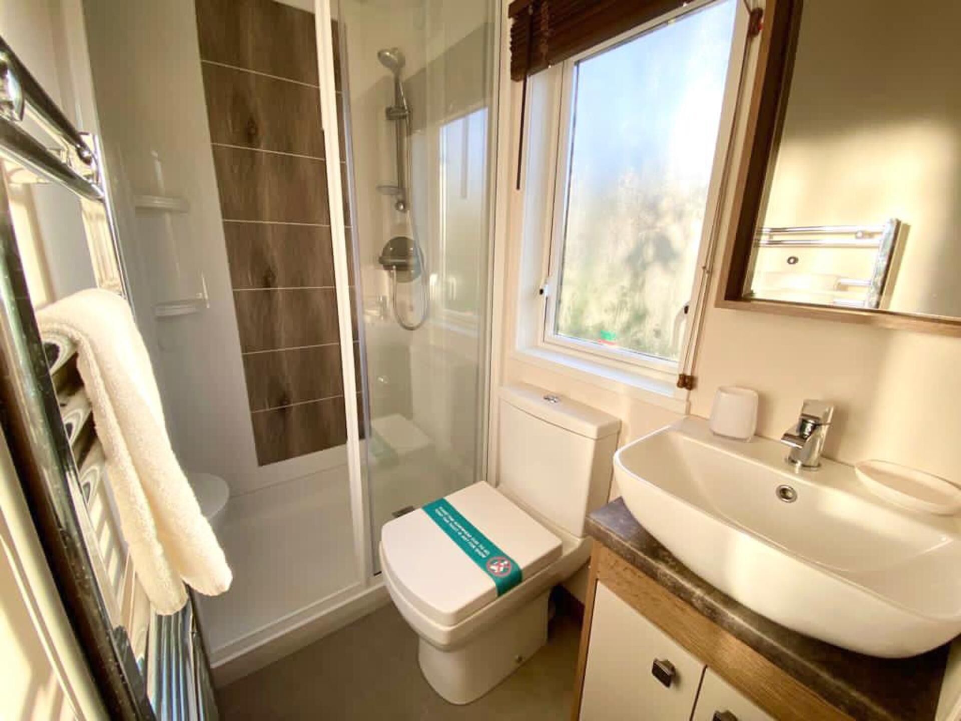 A bathroom with a glass-enclosed shower, a toilet with a green sign, a sink, and a window above the sink.