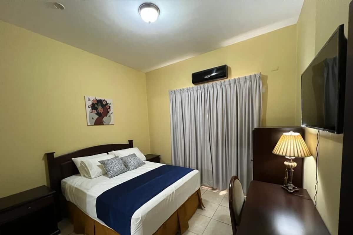 Habitación de hotel con cama, mesita de noche con lámpara, televisor, ventana con cortinas y aire acondicionado instalado en la pared.