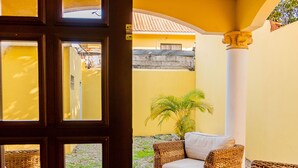 Une porte ouverte donne sur un patio aménagé avec des meubles en osier, un ventilateur de plafond et une vue sur un bâtiment jaune.