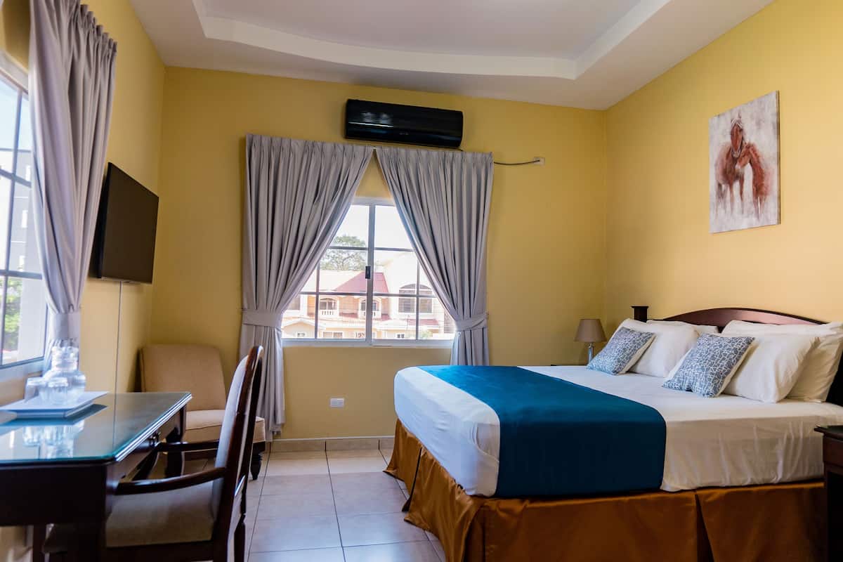 Habitación de hotel con cama, escritorio, silla, ventana con cortinas, aire acondicionado y un cuadro en la pared.