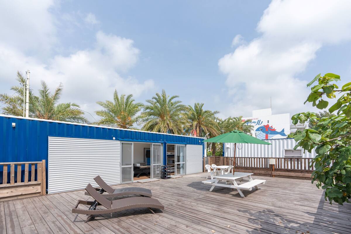 Eine Holzterrasse mit Liegestühlen und einem Tisch, ein blaues Gebäude mit einer Schiebetür und Palmen im Hintergrund.