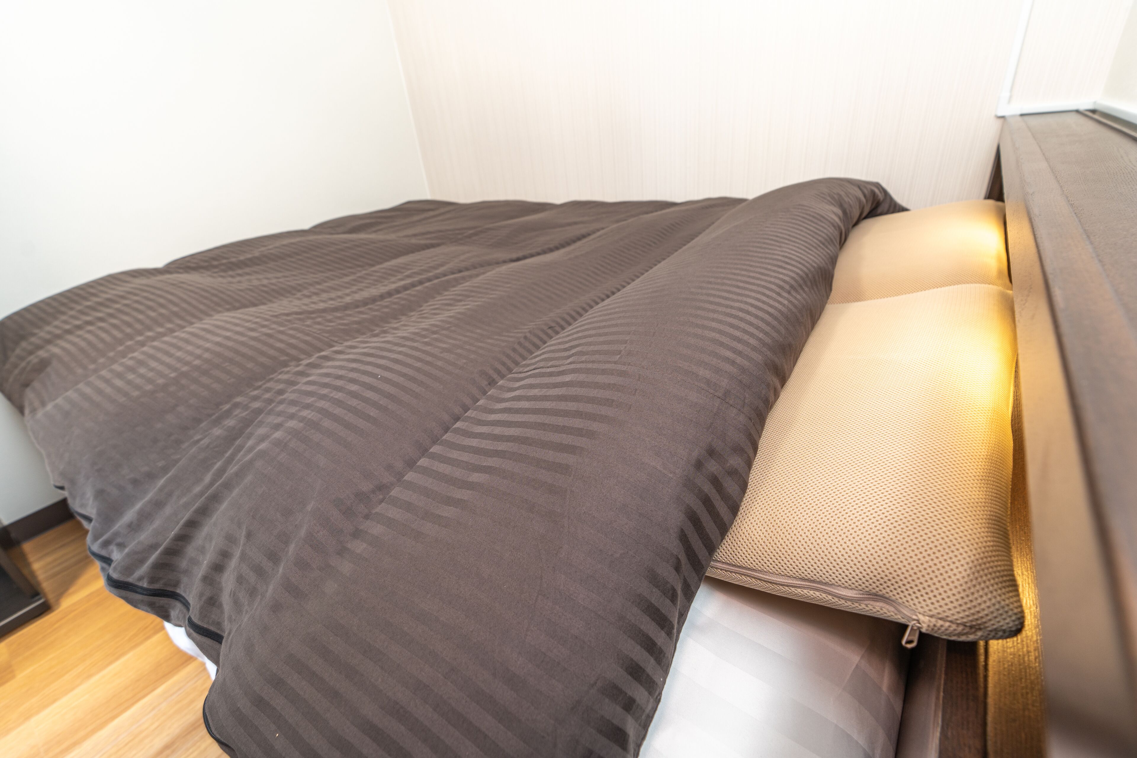 A neatly made bed with a brown blanket and a pillow, a wooden floor, and a white wall.