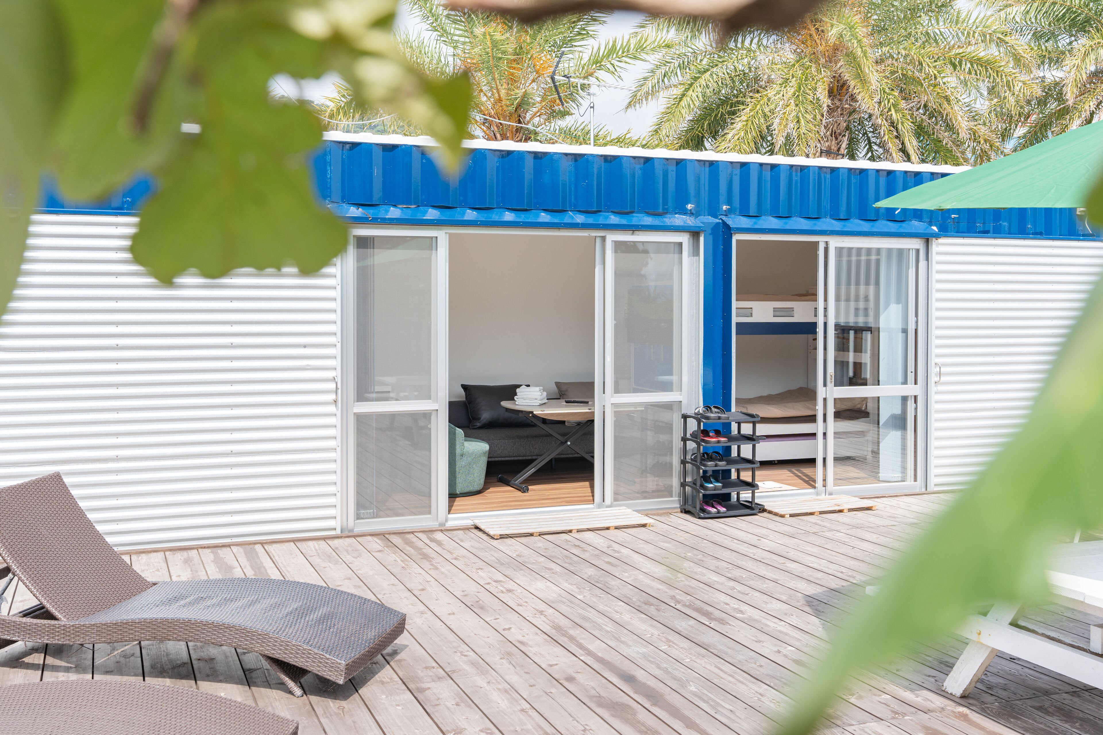 A small, single-story building with a blue roof and white exterior walls. It features a sliding glass door entrance leading to a room with a bed, a table, and a couch. There is a wooden deck outside with a lounge chair and a small table.