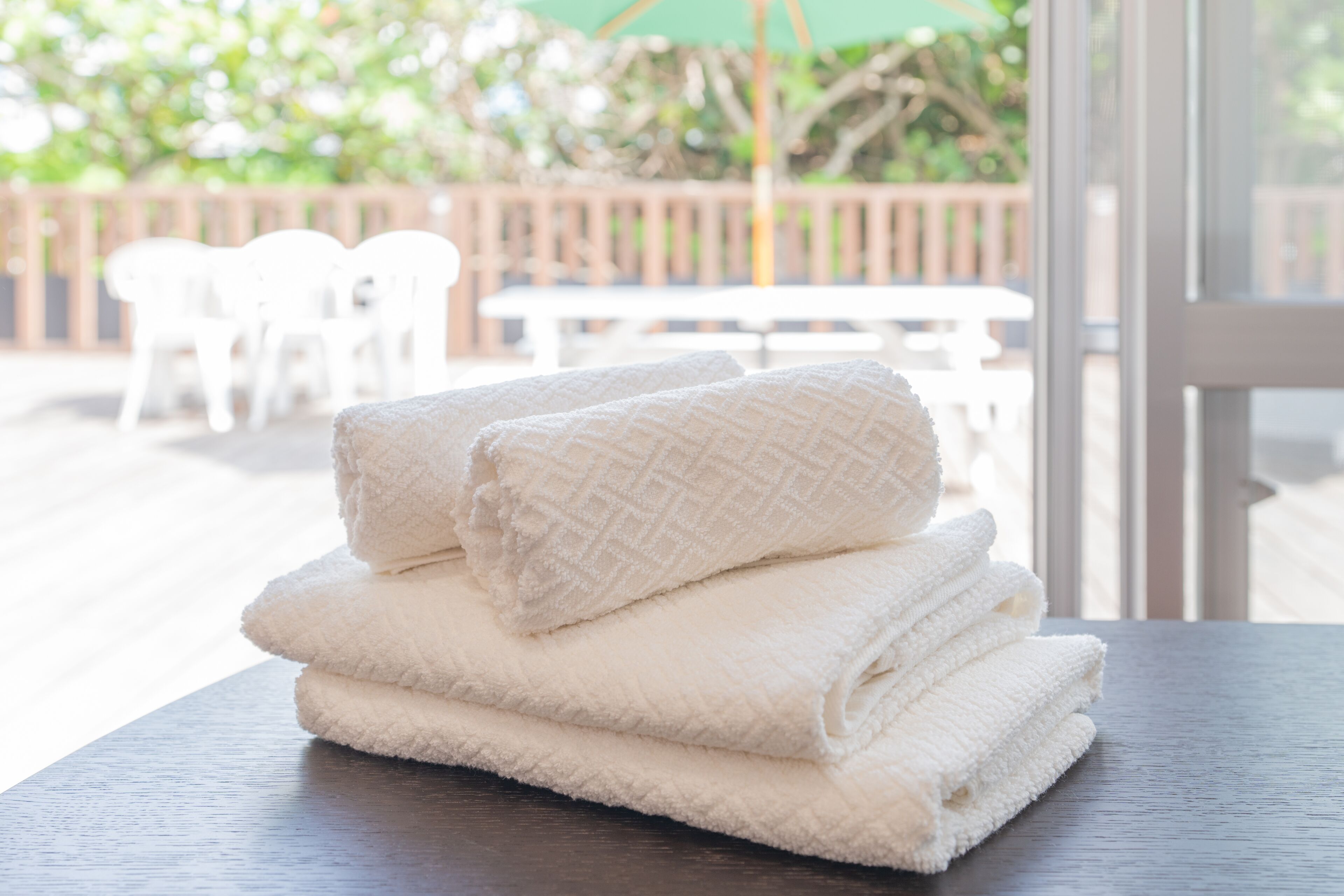 A stack of white towels on a wooden table with a view of outdoor seating and greenery.