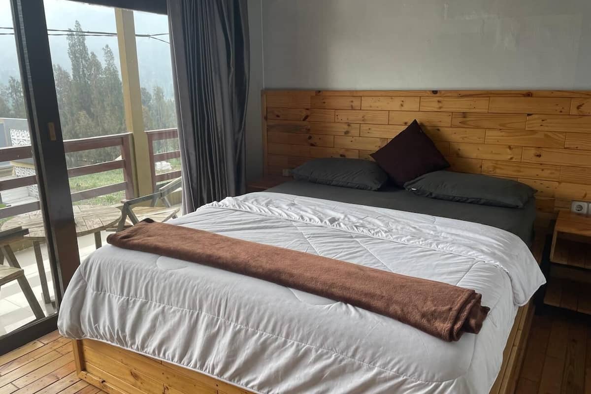 Un lit en bois avec une literie blanche et une serviette marron, une tête de lit en bois et une vue sur un balcon aménagé avec du mobilier d’extérieur.