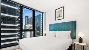 Una habitación de hotel moderna con una cama grande, vistas de edificios urbanos a través de ventanas de pared a techo, un cabecero verde azulado y un cuadro en la pared.
