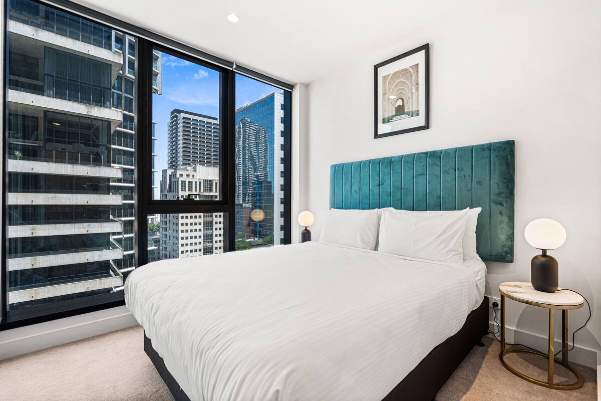 A modern hotel room with a large bed, a view of city buildings through floor-to-ceiling windows, a teal headboard, and a framed picture on the wall.