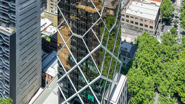 Un edificio moderno de gran altura con una fachada de vidrio geométrica distintiva, rodeado de calles urbanas y áreas verdes.