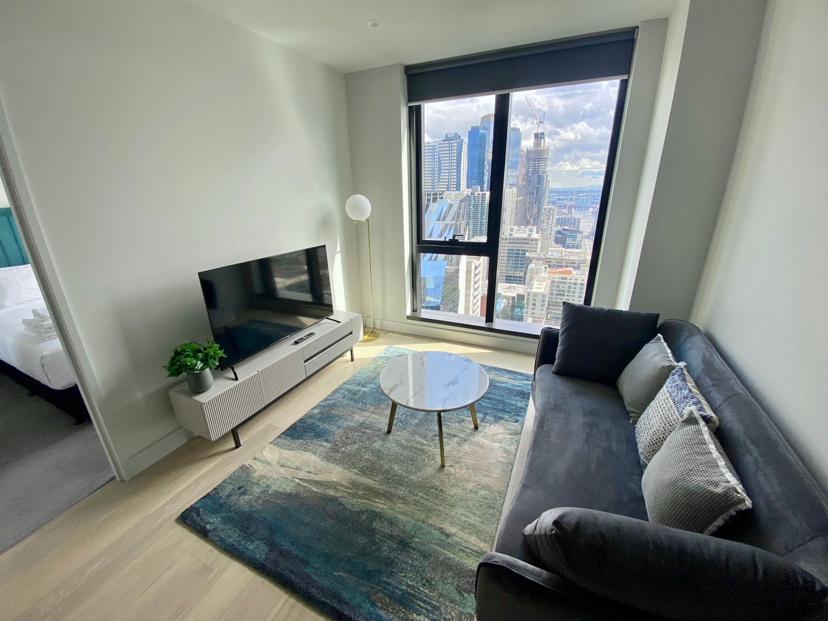 Una sala de estar moderna con un televisor grande, un sofá, una mesa de centro y vistas a un horizonte urbano a través de amplias ventanas.