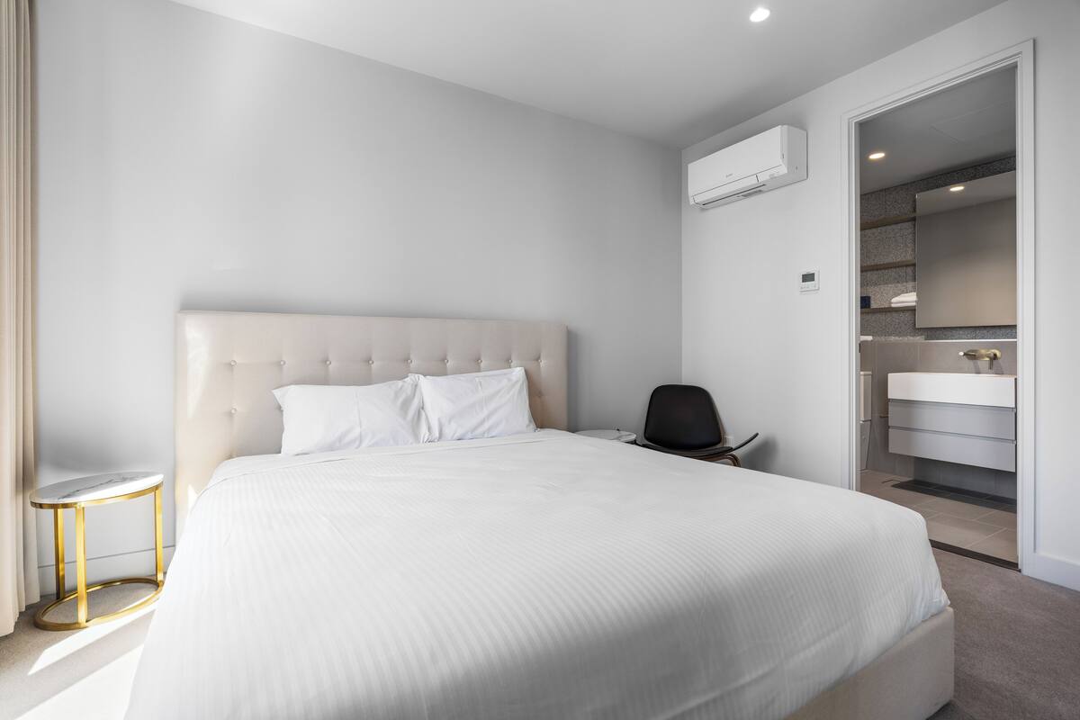 A modern hotel room with a large bed, a bedside table, a chair, a bathroom with a mirror and sink, and an air conditioning unit on the wall.