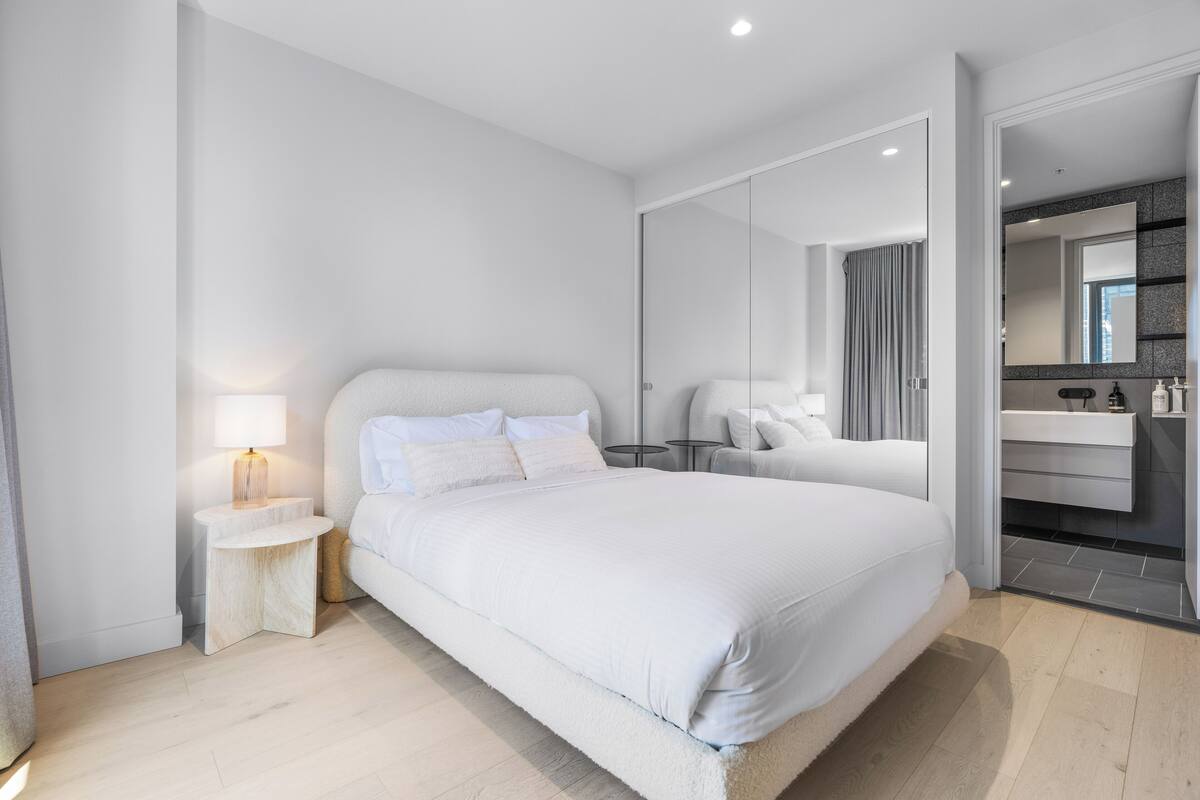 A modern bedroom with a large bed, bedside tables, a nightstand, a mirror, and a bathroom with a sink and mirror.