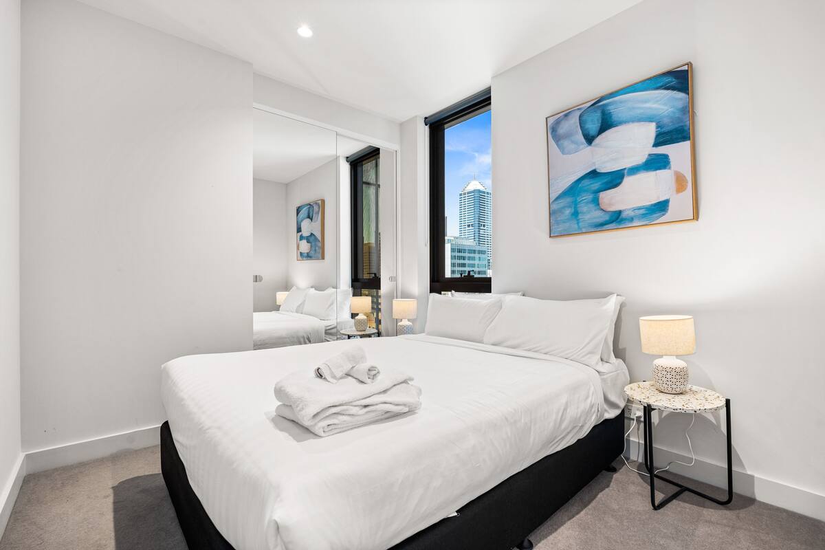A modern hotel room with a large bed, a round side table with a lamp, and a cityscape view through the window.