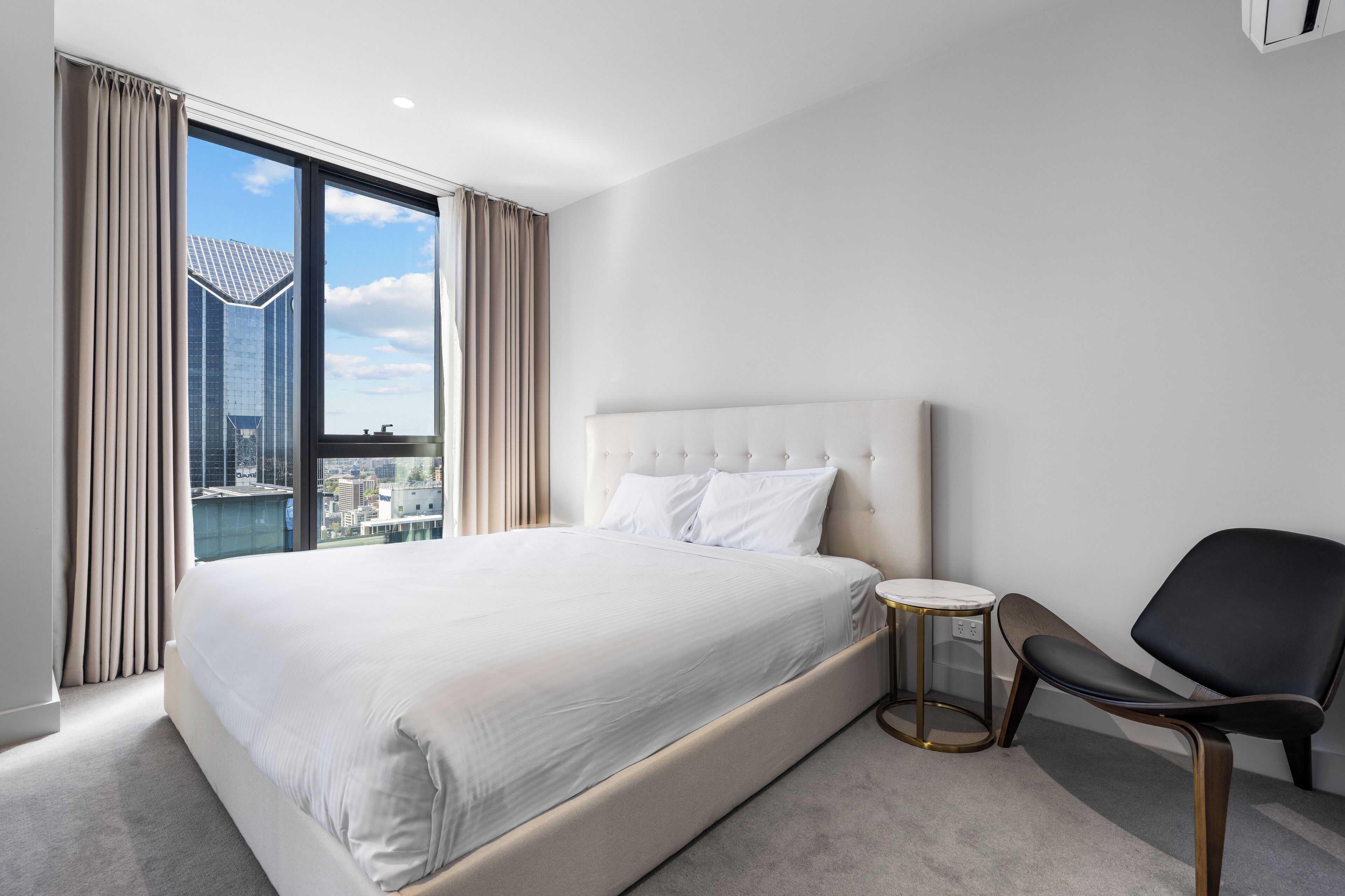 A modern hotel room with a large bed, a round side table, and a view of a cityscape through floor-to-ceiling windows.