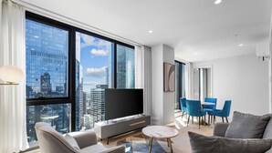 Una sala de estar moderna con un televisor grande, sofás grises, una mesa de centro y un comedor con sillas azules.
