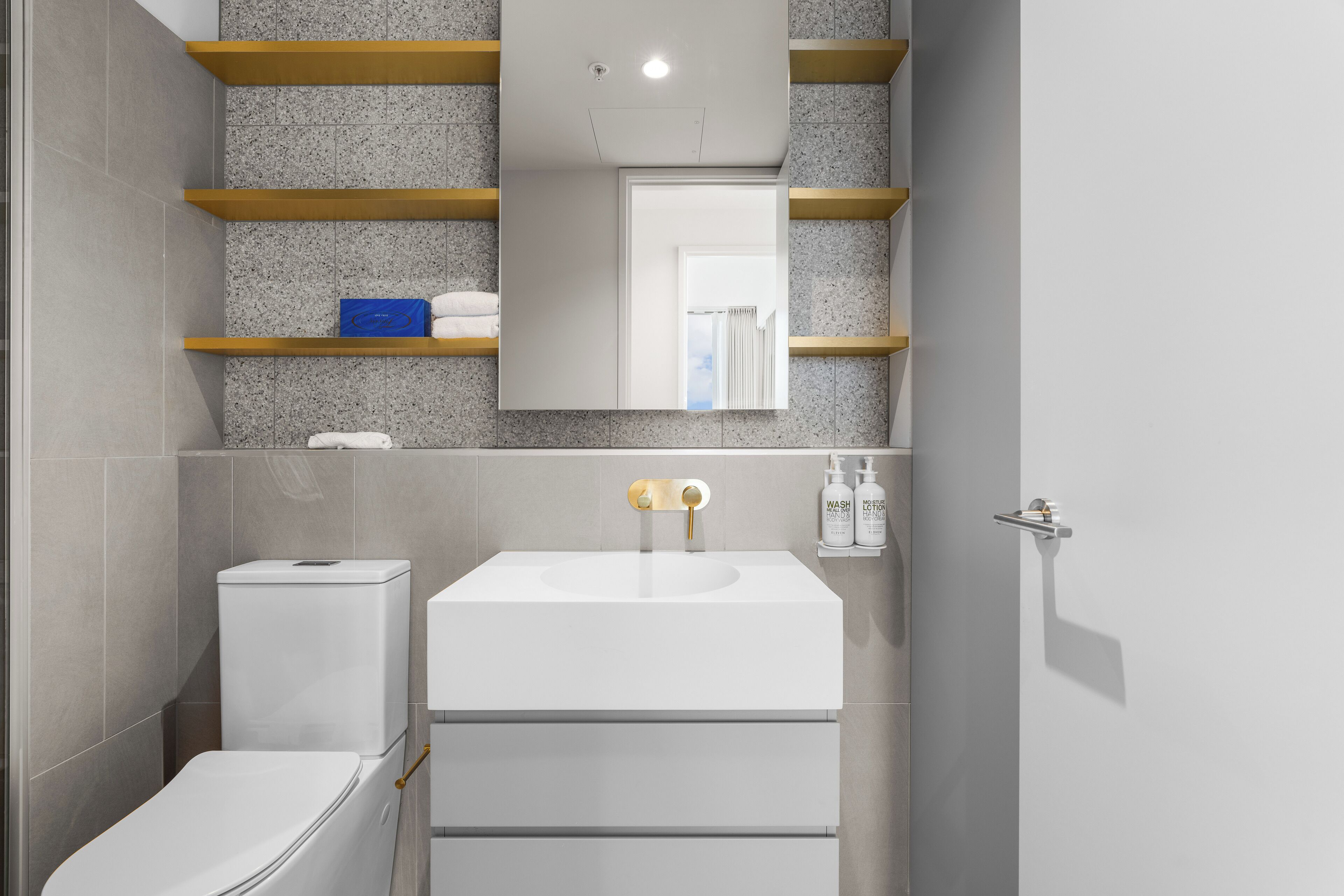 Ein modernes Badezimmer mit einem weißen Waschbecken, einer Toilette und einem Spiegel über dem Waschbecken. Über dem Waschbecken befinden sich Regale und rechts daneben ein Handtuchhalter.