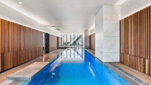 Una piscina interior moderna con suelo de mosaicos azules brillantes, bordeada por paneles de madera y detalles de mármol.