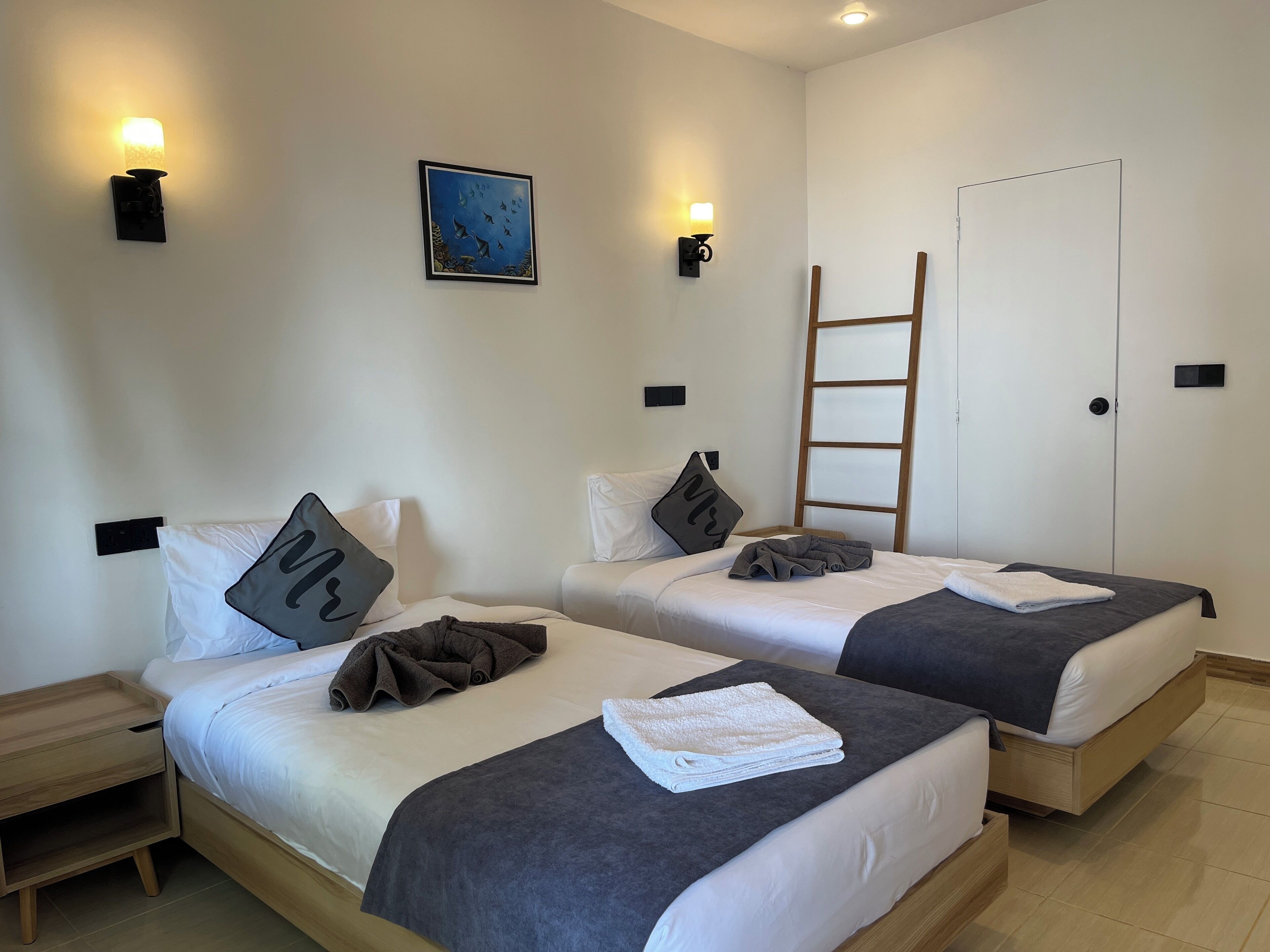 Habitación de hotel con dos camas individuales, cada una con una almohada y una manta. Hay luces fijadas a la pared y un cuadro sobre las camas.