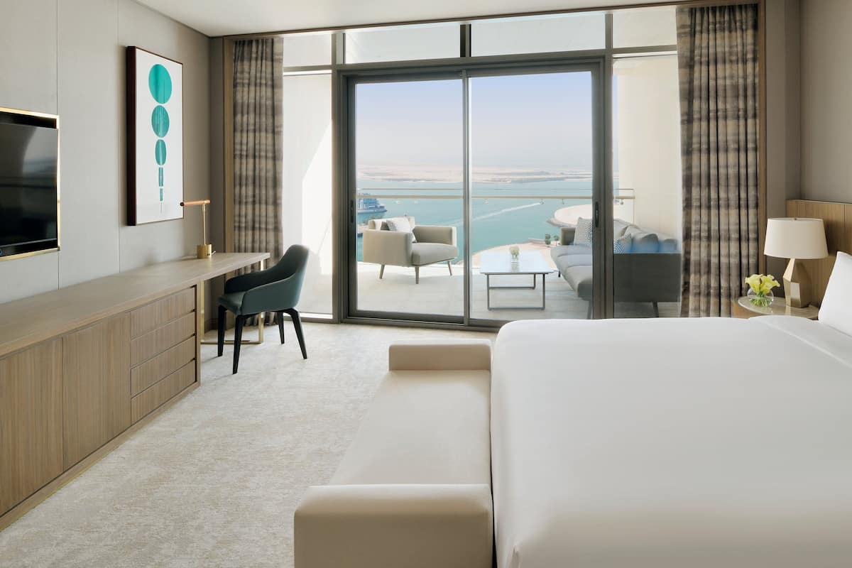 A hotel room with a large bed, a desk with a chair, a television, and a view of the sea through the glass door.