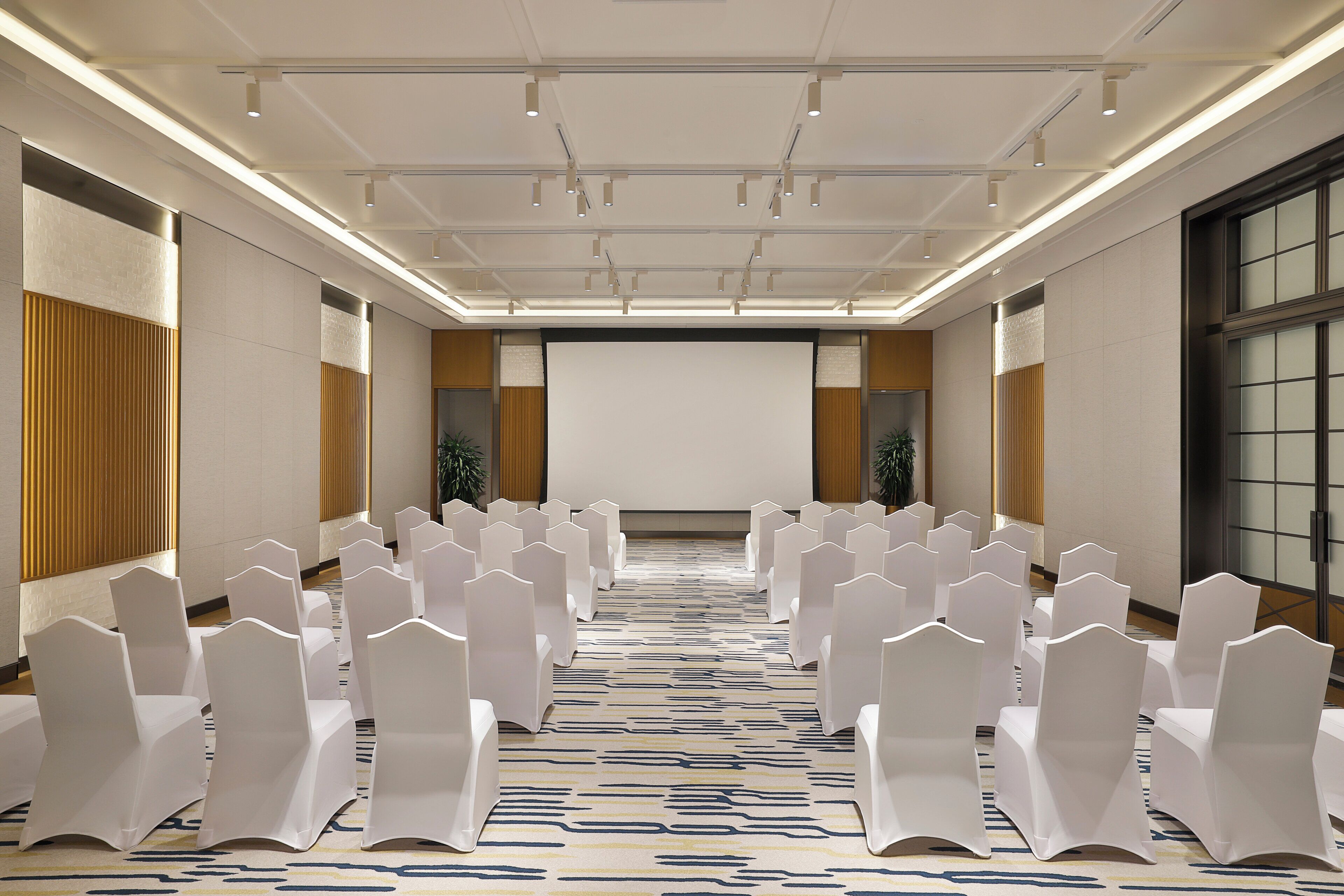 A conference room with rows of white chairs, a projector screen, and a patterned carpet.