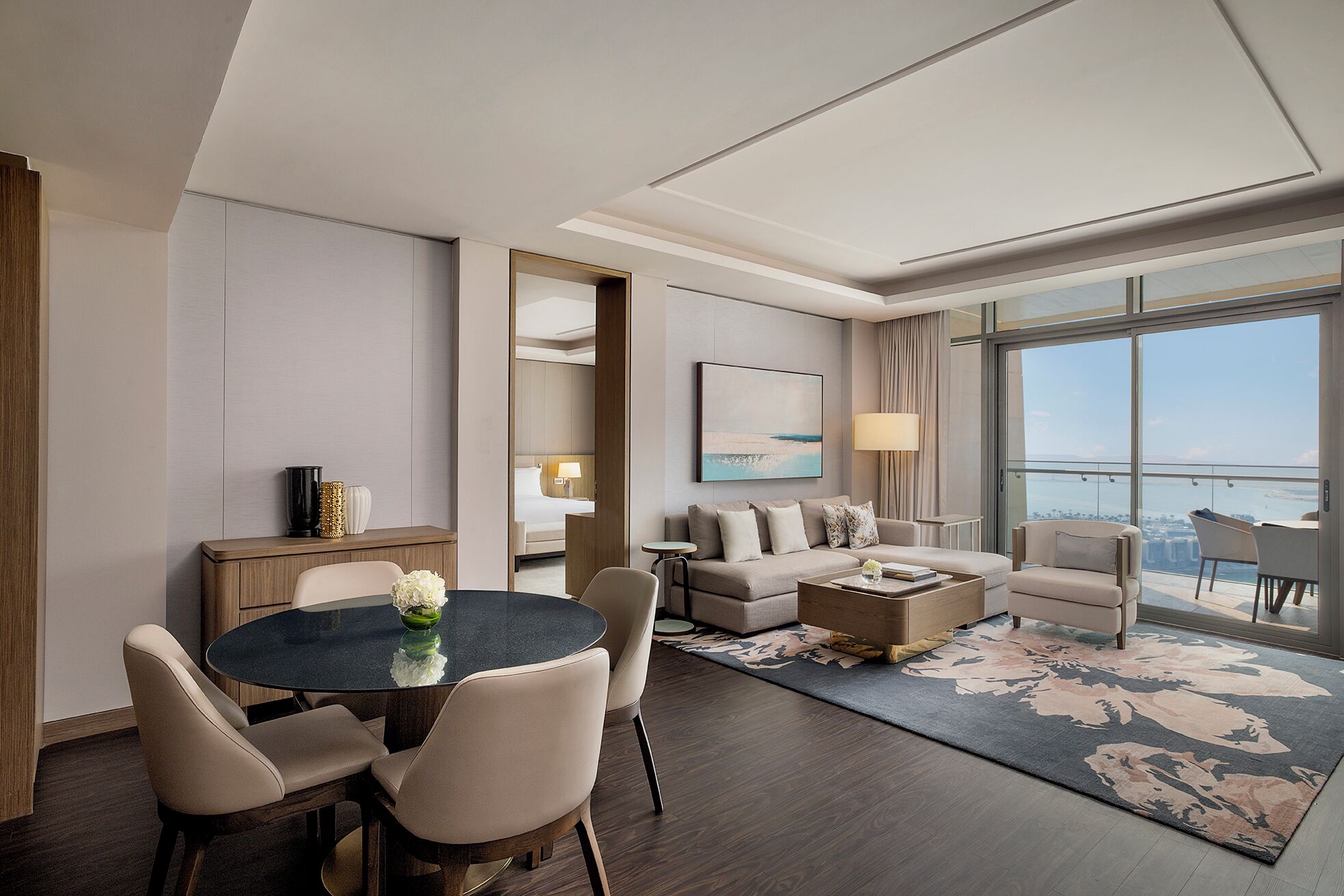 A modern hotel room with a dining area, a sofa set, a large window with a view, and a painting on the wall.