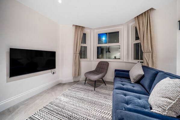 Uma sala de estar moderna com televisão de tela plana, um sofá azul, um tapete estampado e uma janela com cortinas.