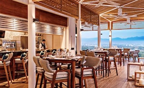 A restaurant with wooden tables and chairs, a bar area, and a view of the city and mountains.