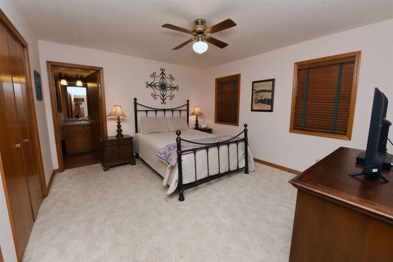 A bedroom with a bed, bedside lamps, a ceiling fan, a desk with a computer, and a window with blinds.