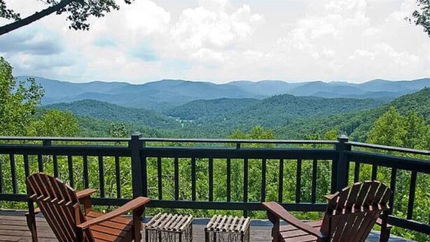 A wooden deck with Adirondack chairs and a small table, overlooking a mountainous landscape.