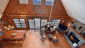 A spacious loft-style living area with a wooden ceiling, a fireplace, a television, and a large window overlooking a deck.