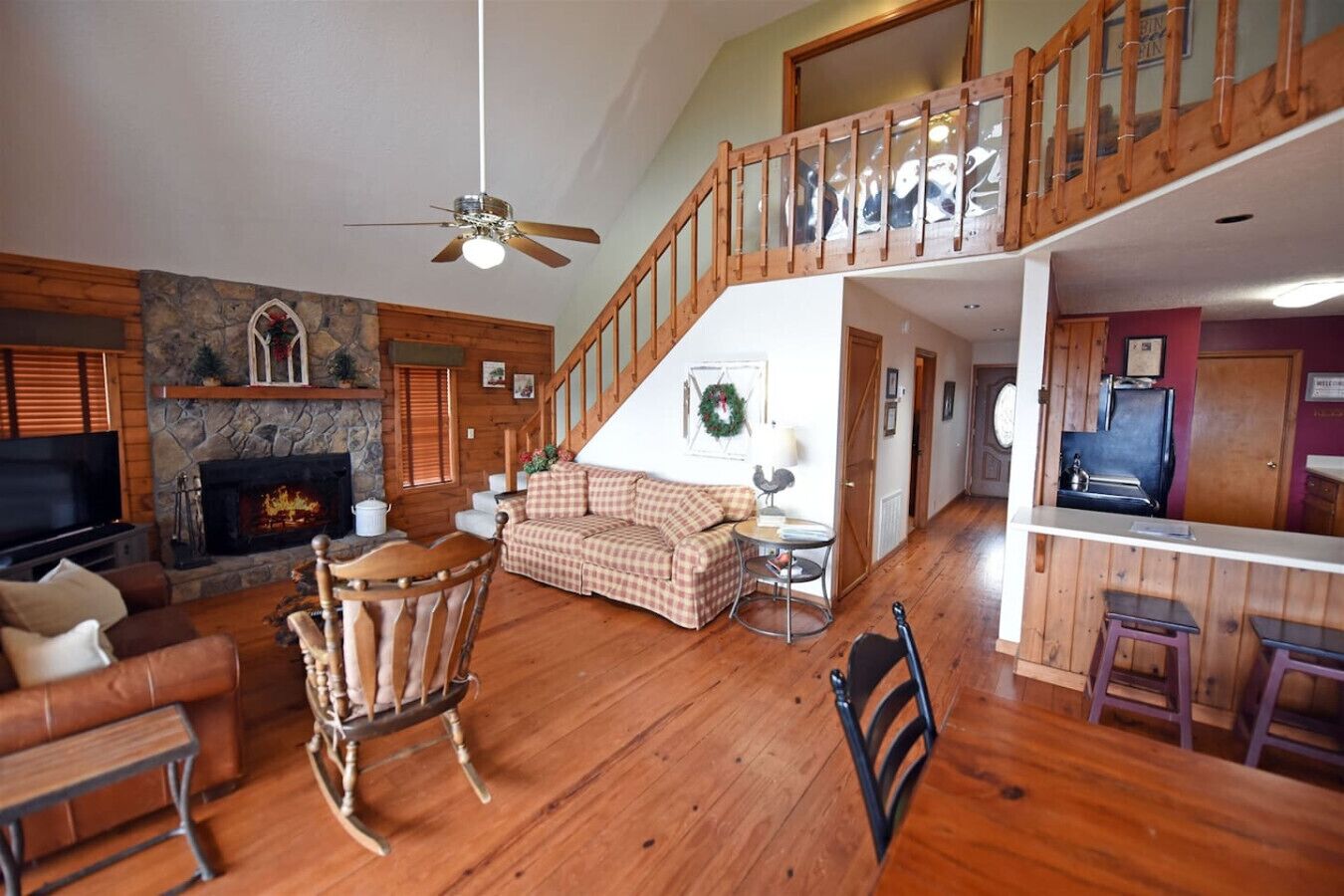 A spacious living area with a fireplace, wooden flooring, a high ceiling fan, a staircase, and a kitchen area.
