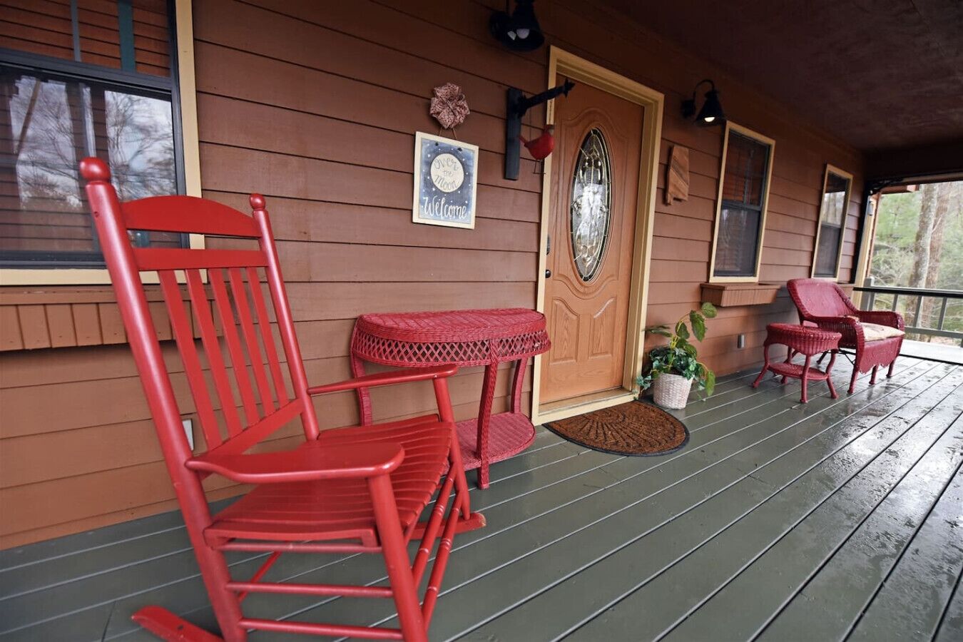 Een houten terras met rode stoelen en een kleine tafel, een welkomstbord en een deur met een decoratief glasraam.