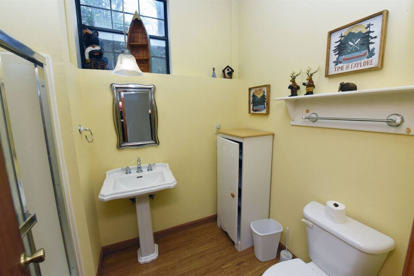 A compact bathroom with a pedestal sink, a toilet, a small trash bin, and a mirror.