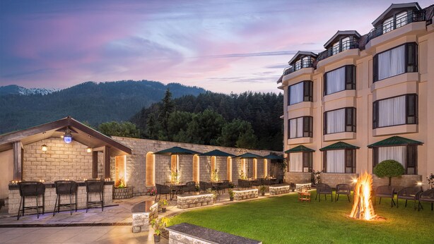 A hotel with outdoor seating, a fire pit, and a mountain backdrop. - Kanishka, Manali- AM Hotel Kollection (Manali)