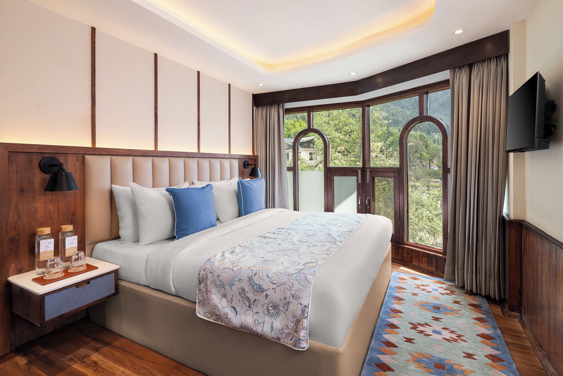 A modern hotel room with a large bed, a nightstand with a lamp, a TV, and a window with curtains.