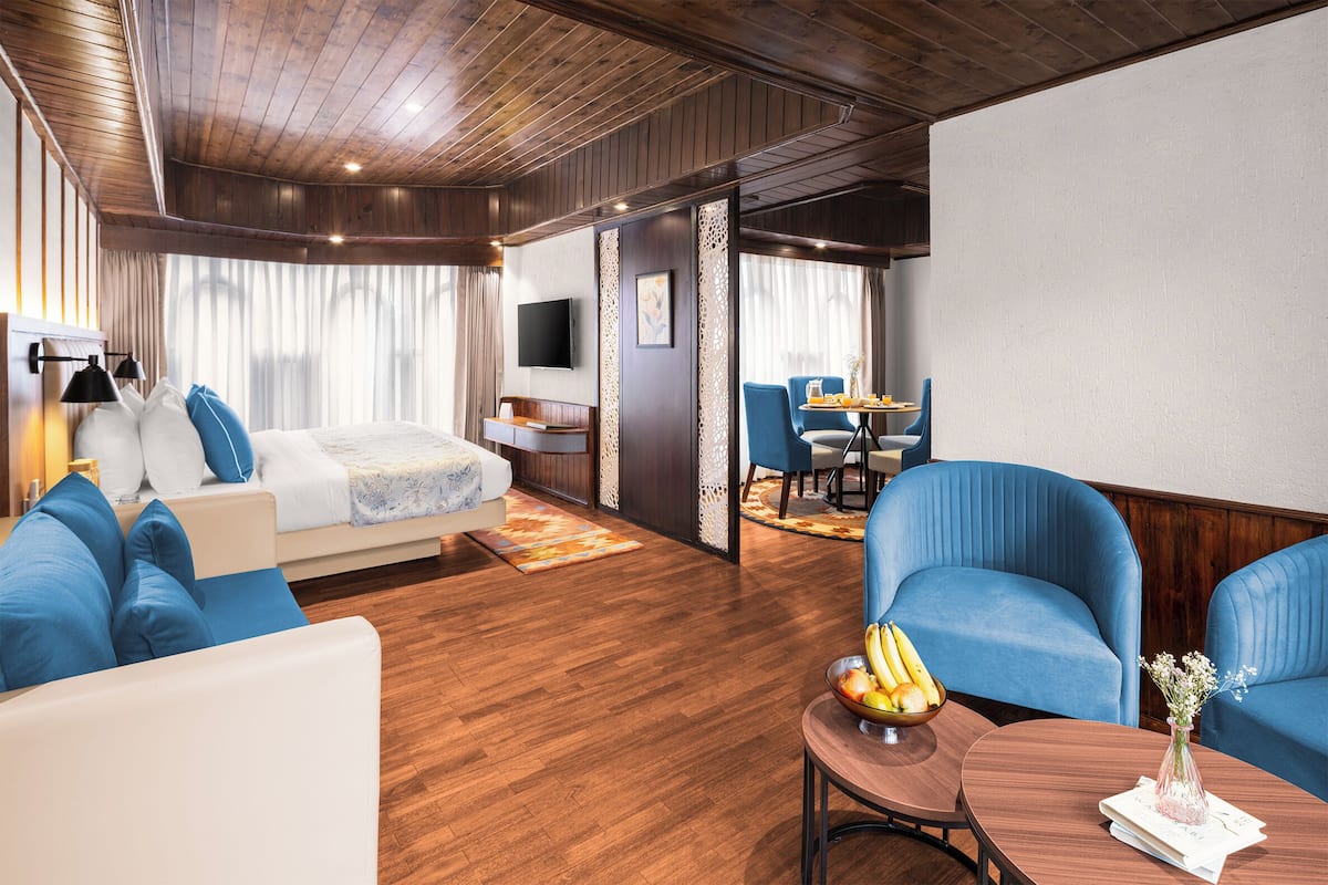 A hotel room with a wooden ceiling, a bed with blue and white bedding, a seating area with blue chairs, a dining table with a bowl of fruit, and a TV mounted on the wall.