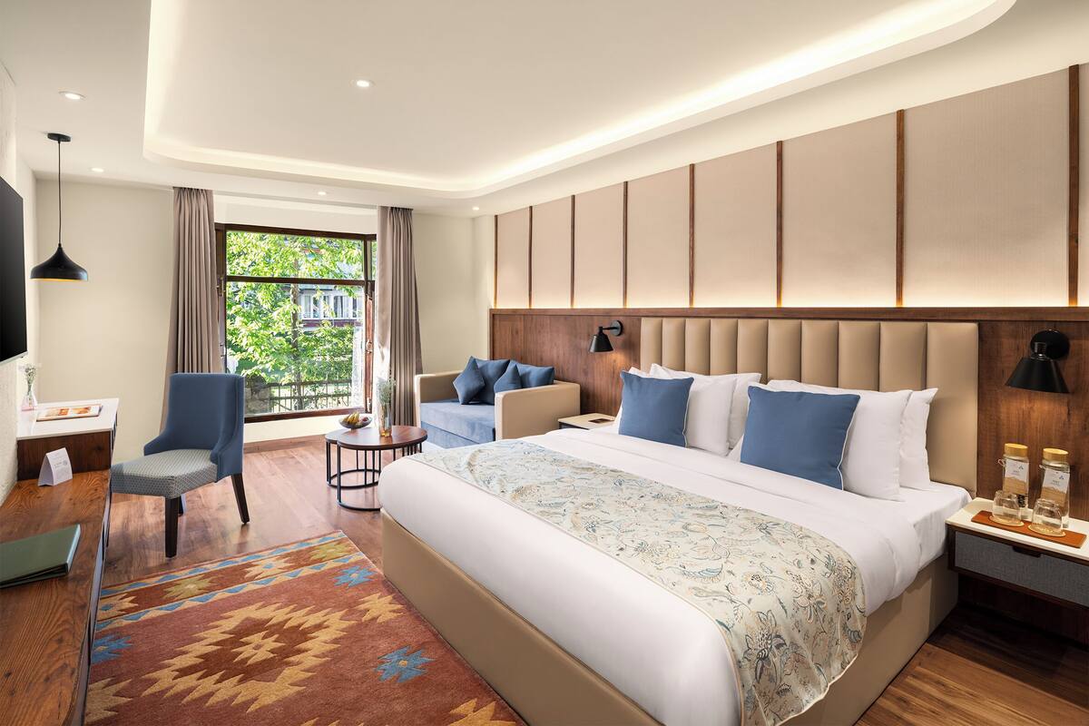 A modern hotel room with a large bed, a desk, a chair, and a view of greenery outside.