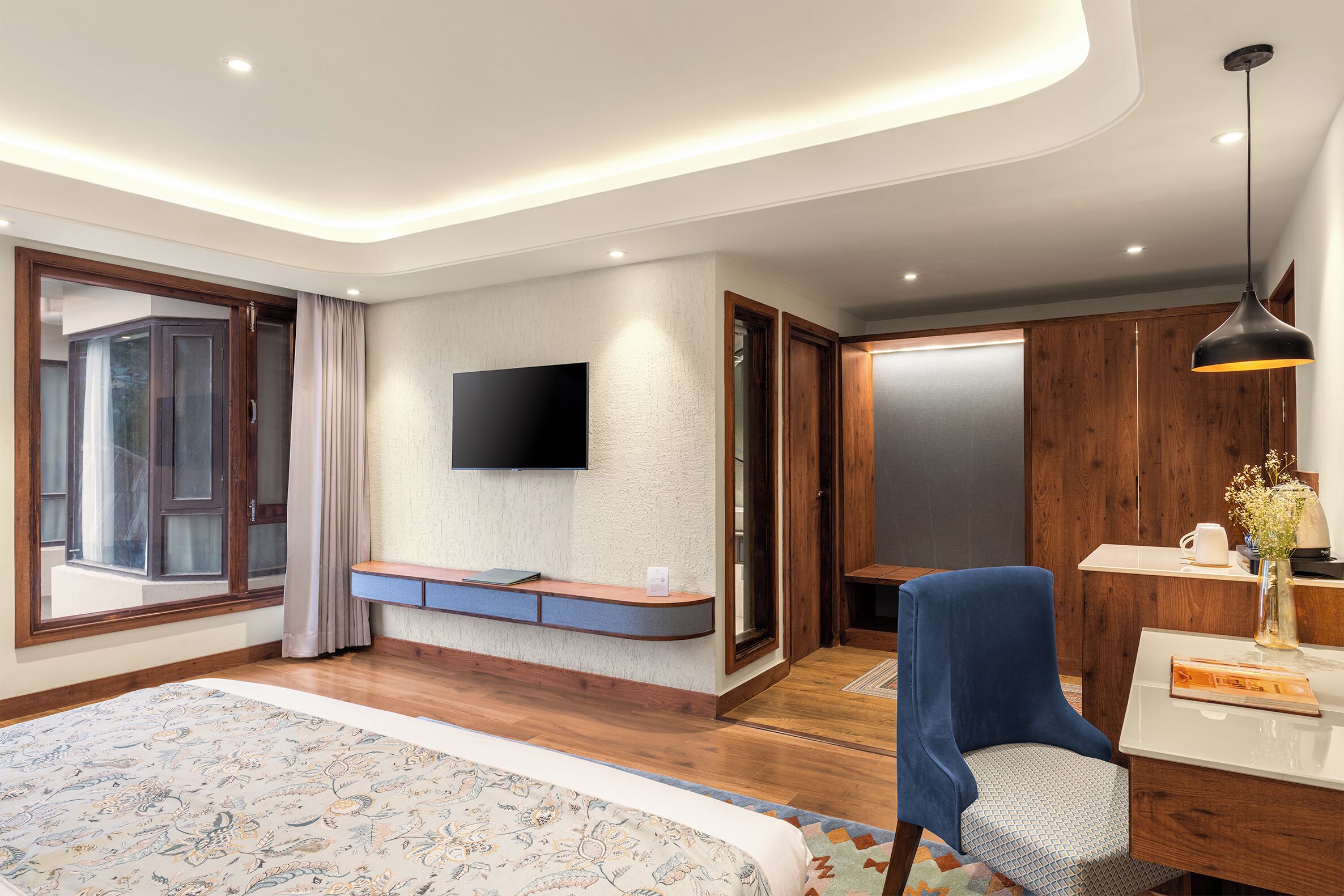 A modern hotel room with a large bed, a desk, a chair, a television, and a wardrobe.