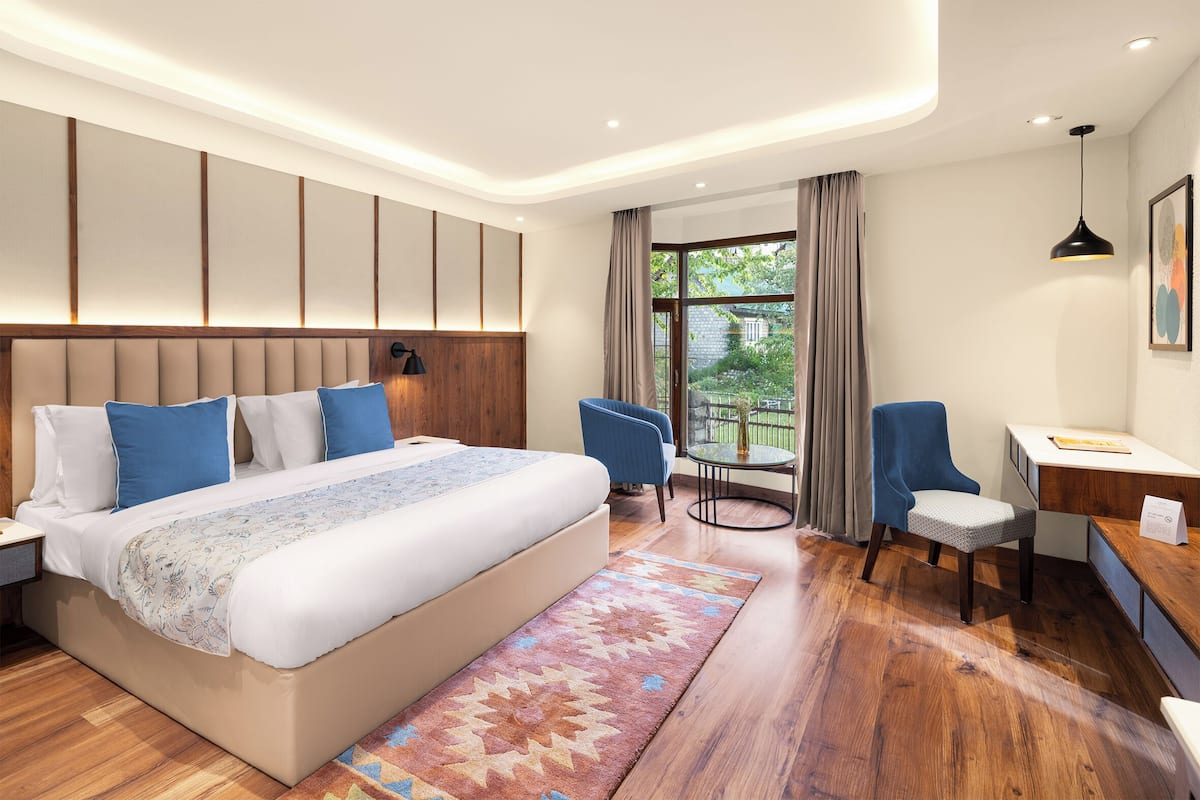 A modern hotel room with a large bed, two chairs, a desk, and a view of greenery outside.