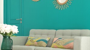 Una pared azul verdoso con espejos decorativos y plantas, un sofá gris con cojines coloridos y una mesa de centro de madera con un jarrón de flores.