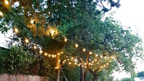 Un área de estar al aire libre con sillas azules y una terraza de madera, rodeada de árboles y luces colgantes.