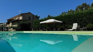 Une piscine à l’eau d’un bleu limpide, entourée d’une clôture. On aperçoit, en arrière-plan, un bâtiment en pierre avec une cheminée, et un parasol blanc qui offre de l’ombre.