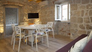A stone-walled room with a round dining table, white chairs, a flat-screen TV, and a window with open shutters. - Villa Veli Varos, 4 bedrooms, near Diocletian pallace, Split old town (Split)
