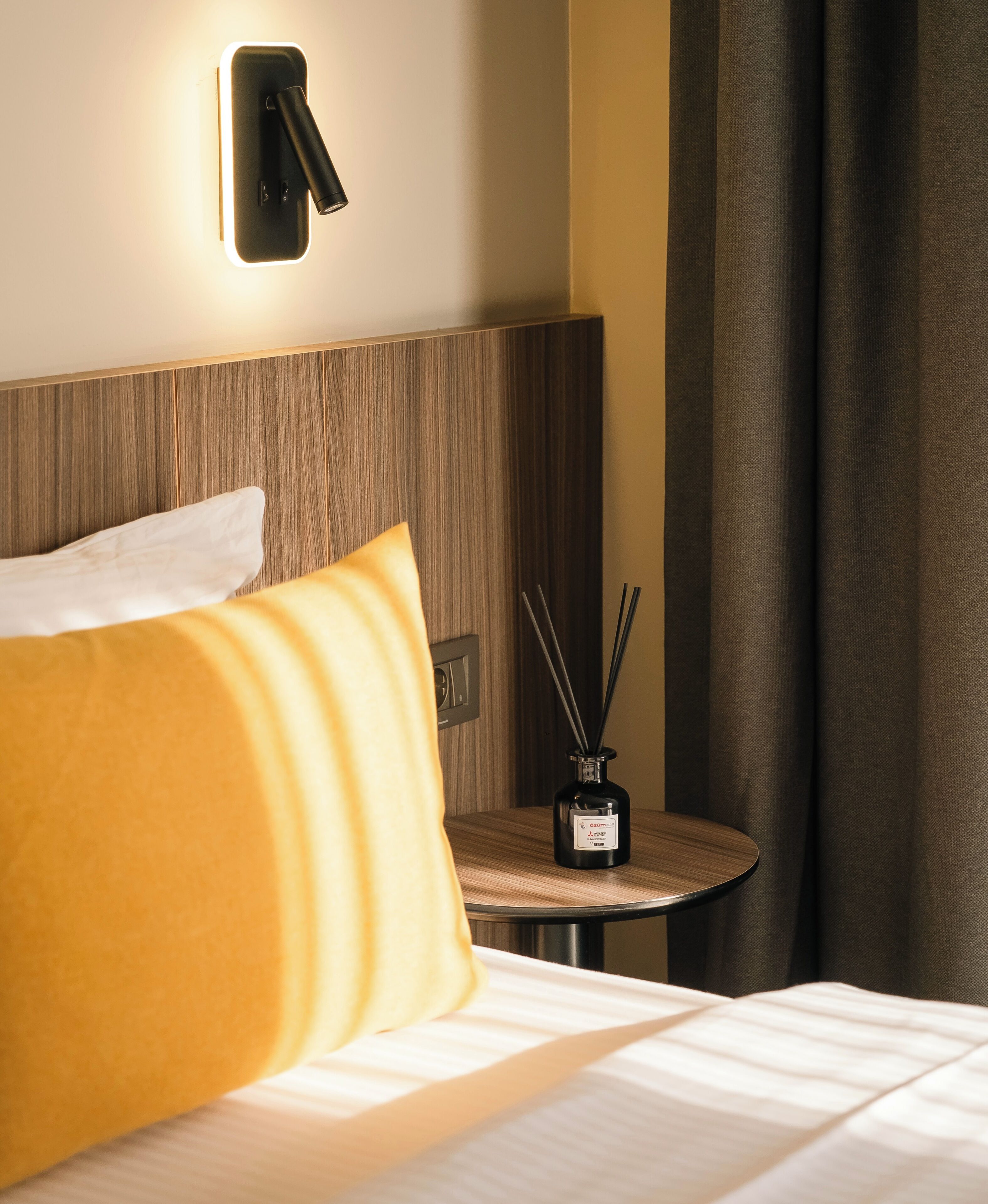 Une chambre d’hôtel comprenant un lit, une table de chevet, une lampe fixée au mur et une table sur laquelle se trouve un diffuseur d’ambiance à bâtonnets.