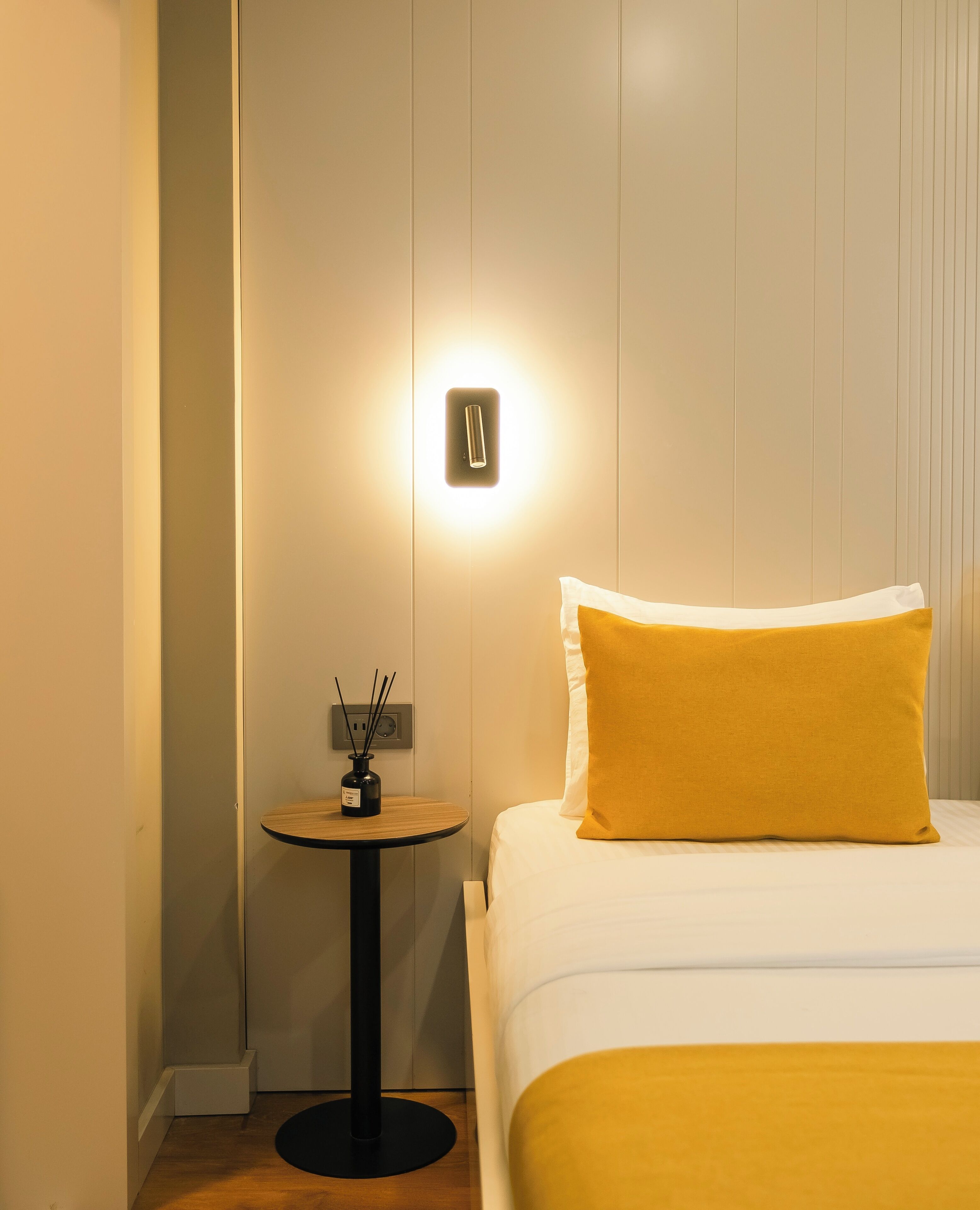 A hotel room with a bed, a bedside table with a lamp, and a wall-mounted light.