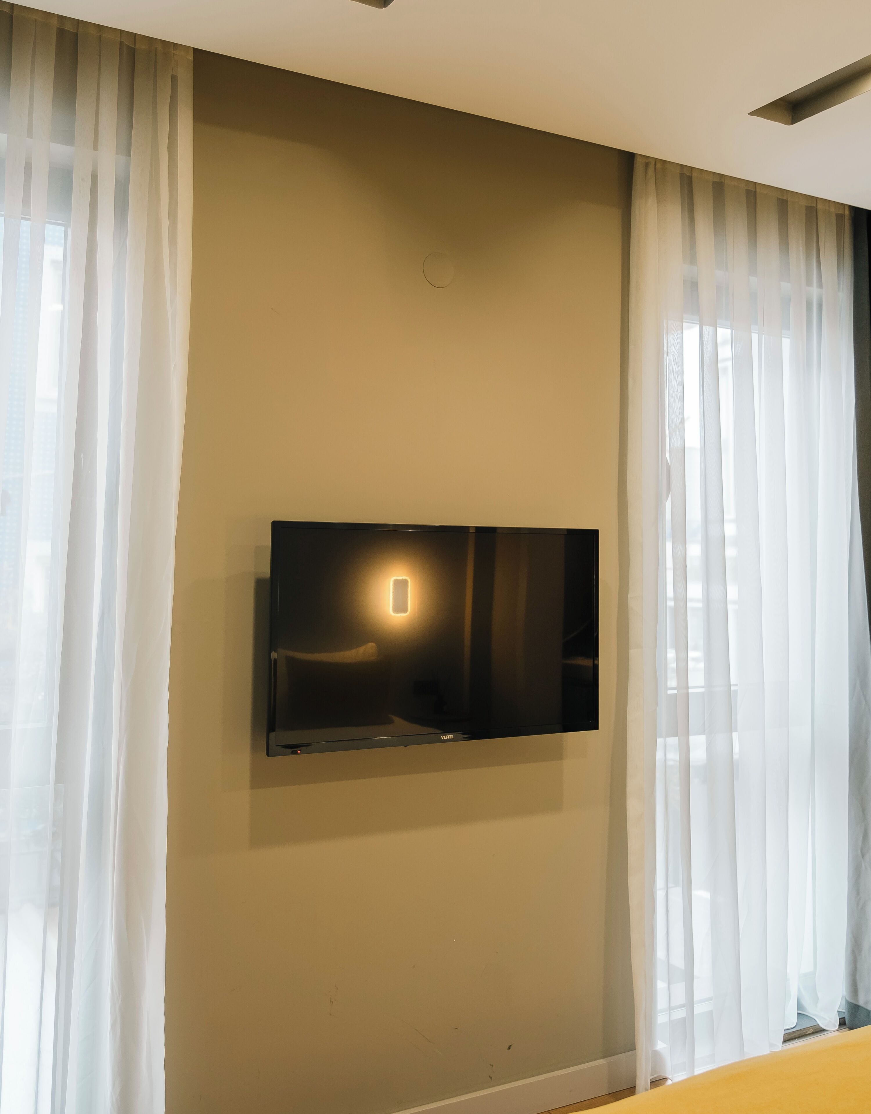 Una habitación con un televisor de pantalla plana montado en la pared, cortinas translúcidas en las ventanas y una pared beige lisa.