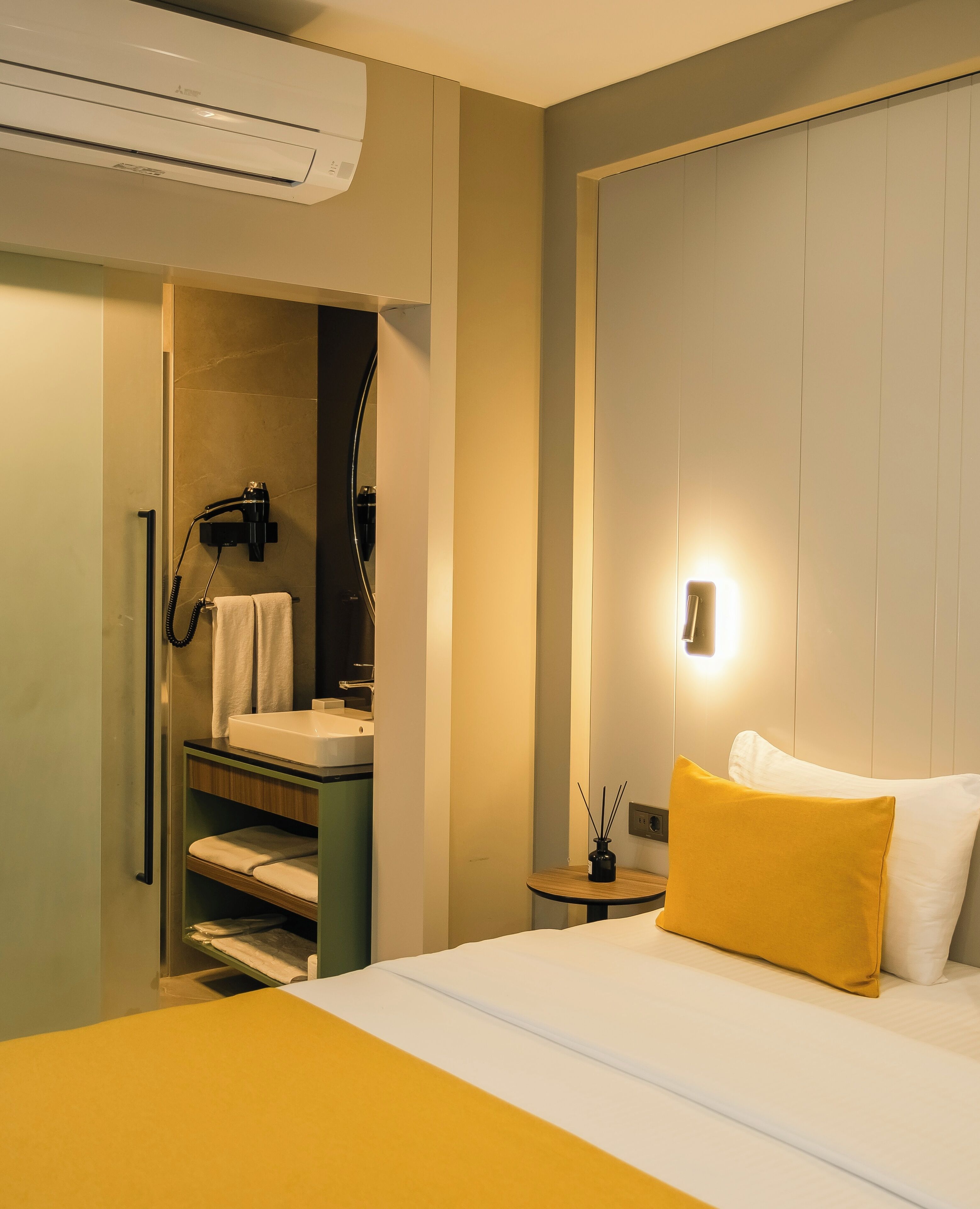 A hotel room with a bed, a bedside table with a lamp, a bathroom with a sink and a hairdryer, and an air conditioning unit.