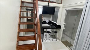 Quarto compacto com escada de madeira que leva a um mezanino com área de dormir. Uma cozinha compacta com fogão e pia é visível, assim como uma televisão montada sobre o balcão.