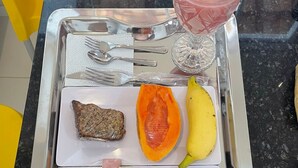 Uma bandeja com alimentos, incluindo bife, ovos, presunto, pimentão e uma banana, além de um copo de bebida rosa.
