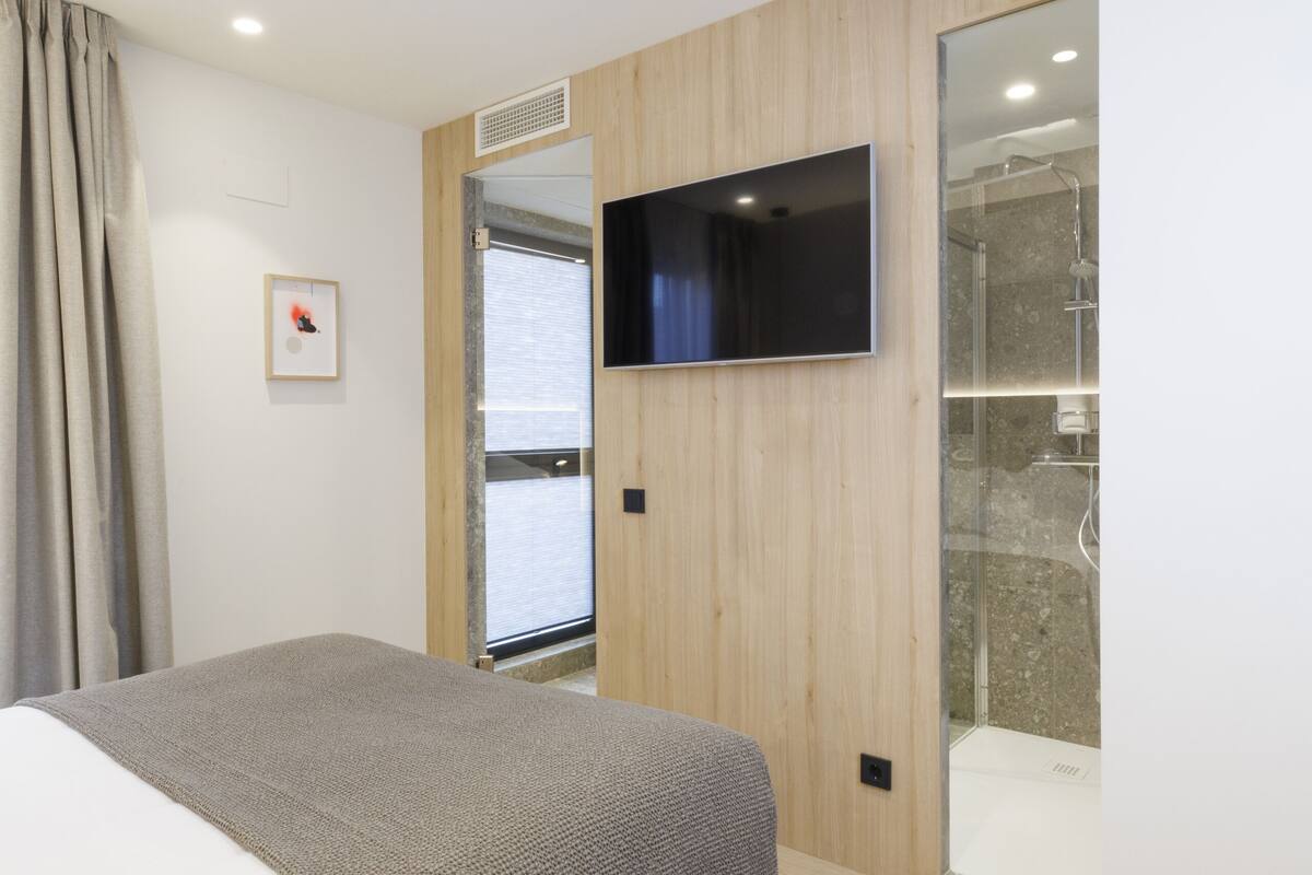 Una habitación de hotel moderna con una cama grande, un televisor de pantalla plana montado en la pared, zona de ducha y diseño con paneles de madera.