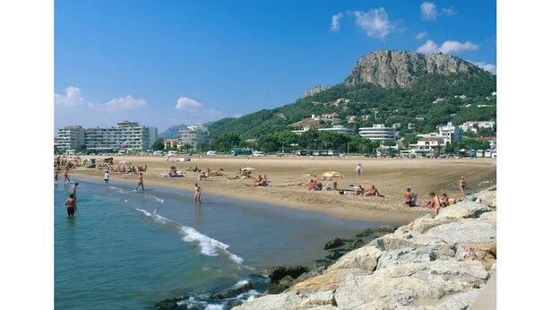 Una playa con gente tomando el sol y nadando, un litoral rocoso y edificios al fondo.
